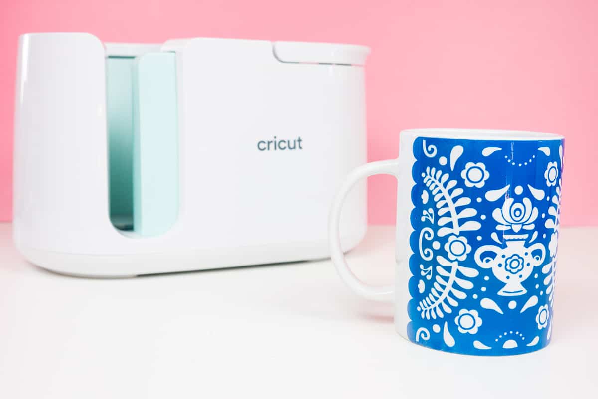 Mug with blue flower pattern in front of Cricut Mug Press on pink background.