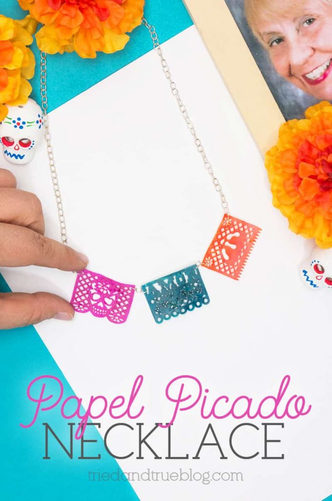 Hand holding Dia de los Muertos Papel Picado Necklace surrounded by holiday artifacts.