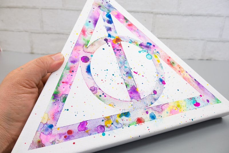 Close up of a hand holding a triangular canvas with watercolor Deathly Hallows logo.