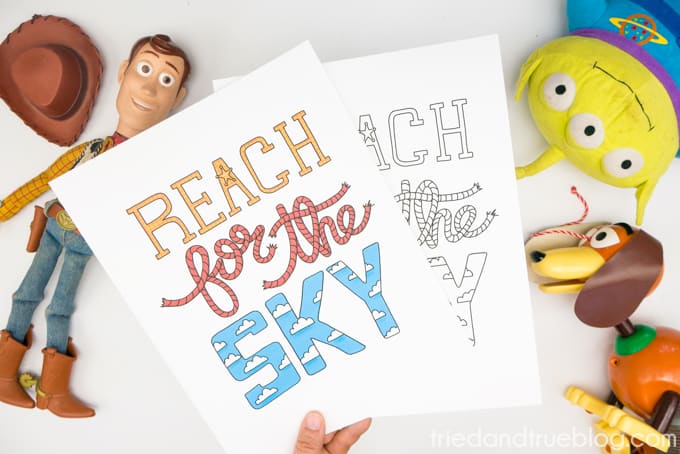 Hand holding two Toy Story "Reach for the Sky" Free Printables surrounded by toys.